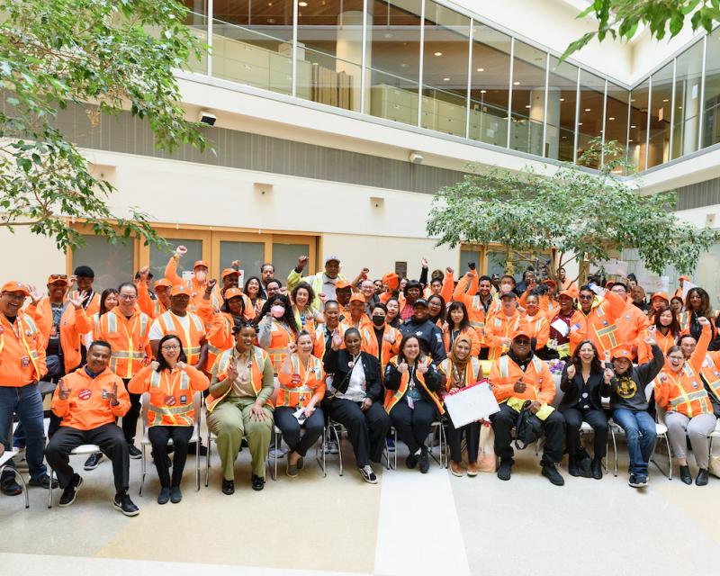 A group of SFMTA staff wearing orange and smile and wave to a camera.