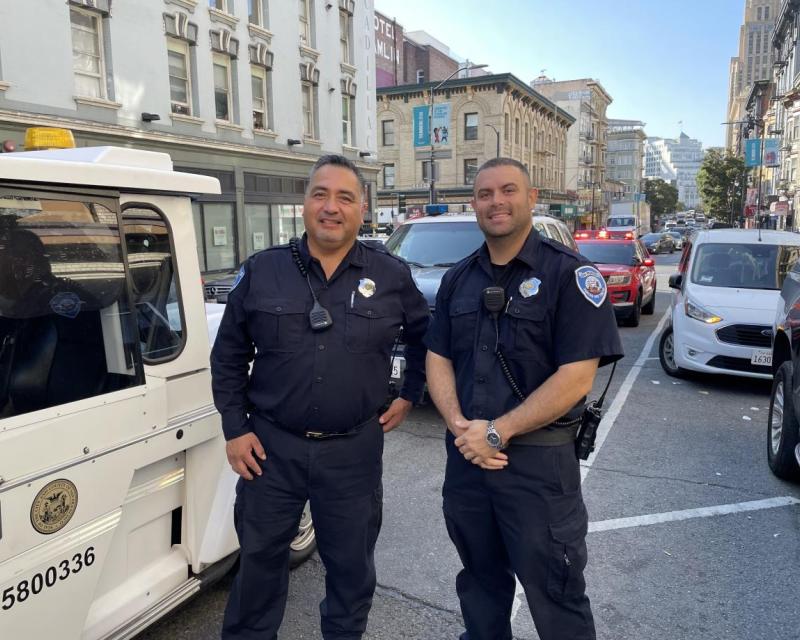 Two Parking Control Officers stand on the side of a busy street with cars and buildings in the background.