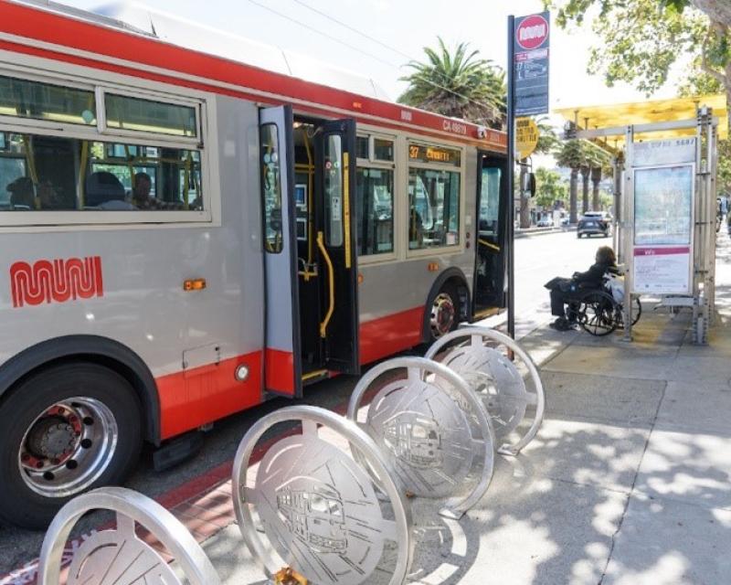 Muni bus with doors open waiting at a bus stop and picking up a person in a wheelchair.