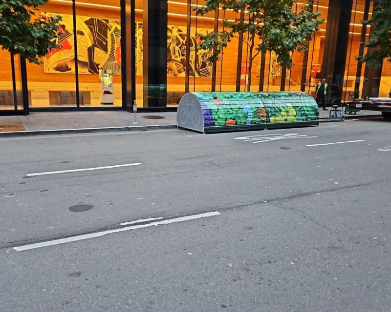 A decorated storage unit is seen on the ground alongside the sidewalk in between two trees.