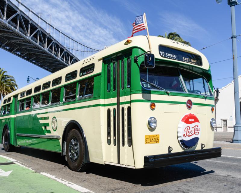 A vintage Muni bus that's green and cream in color passing under a bridge 