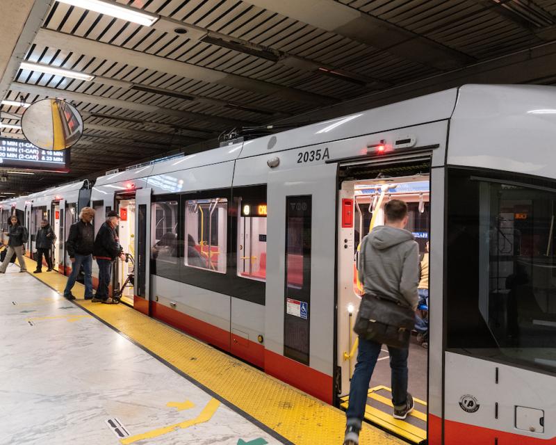 Image shows a train paused at a station platform. Some customers exit as others enter.