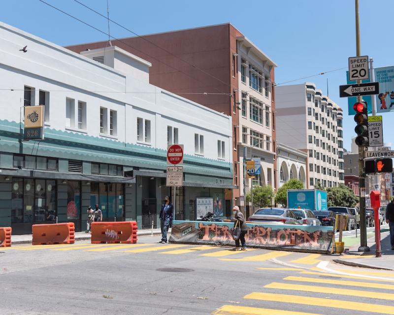 Image shows a person using a crosswalk in front of barricades that block off the street for the Golden Gate Greenway. 