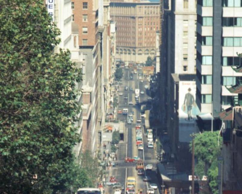 Cable Car 53 climbs Nob Hill on California Street with the Bay Bridge in the background and riders holding onto the poles.