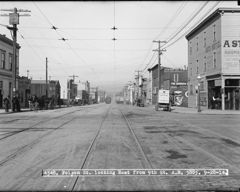 Black and white photo of Folsom Street in wide view, lined by commercial buildings with horses and carriages parked out front.