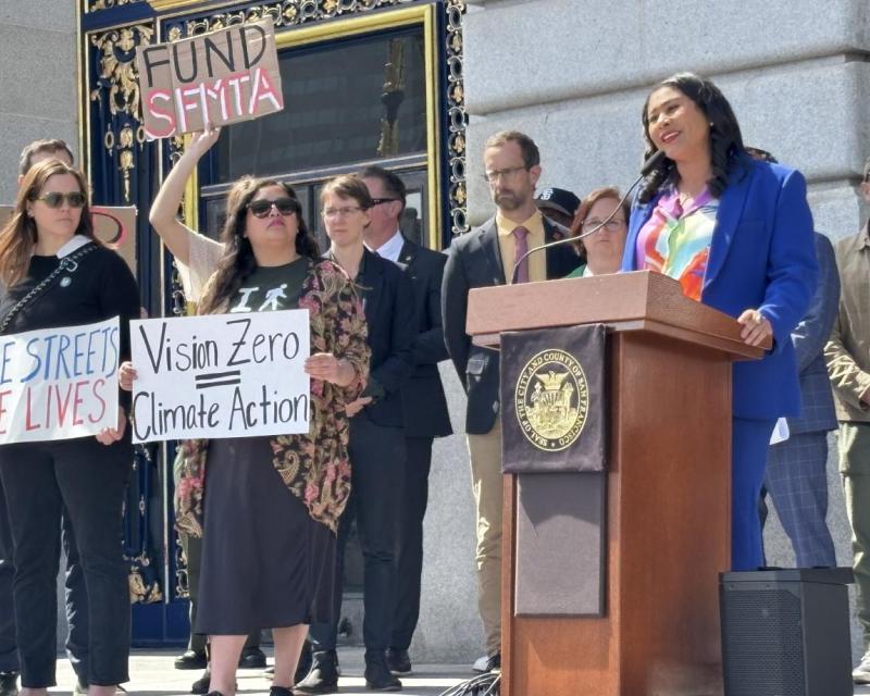 Mayor Breed speaks at a podium in front of city hall. People stand near her with signs that say Fund SFMTA and Vision Zero.