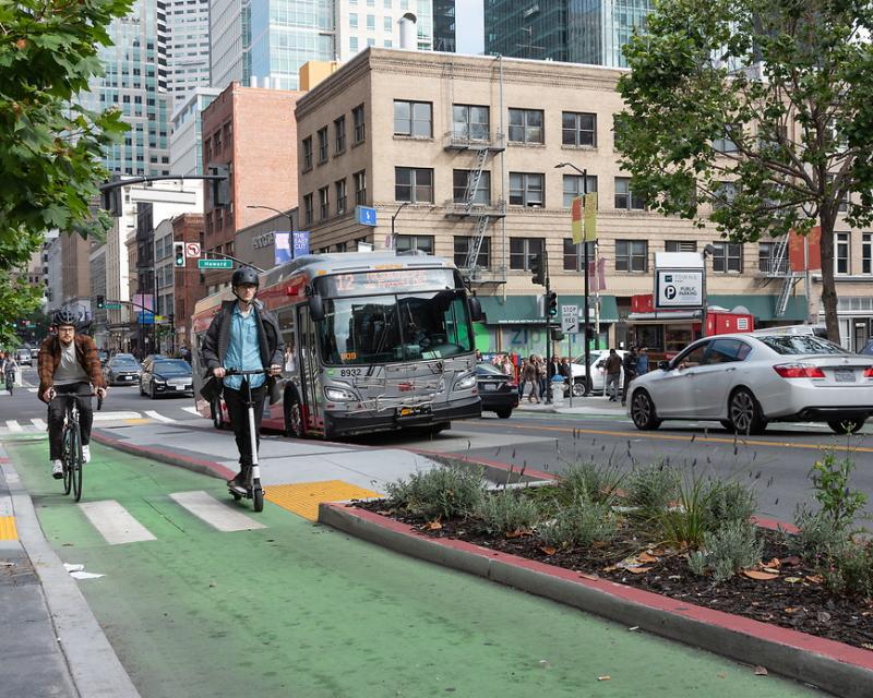 A person rides a scooter in the protected 2nd Street bike lane alongside a cyclist. A Muni bus is visible in the background.