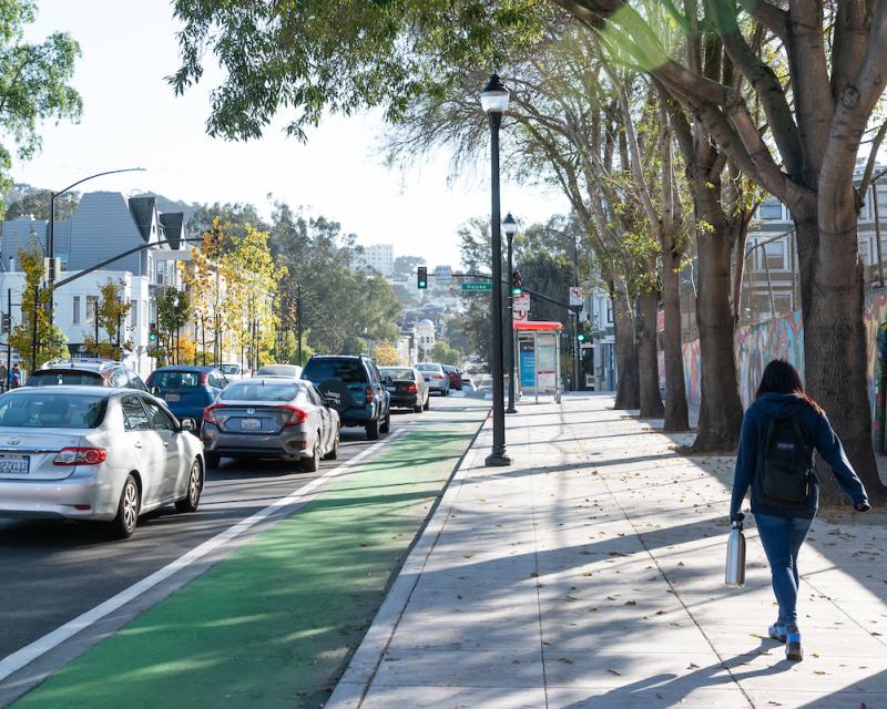 Woman walks on a sidewalk toward a bus shelter. On her left is a green bike lane beside lanes of cars.