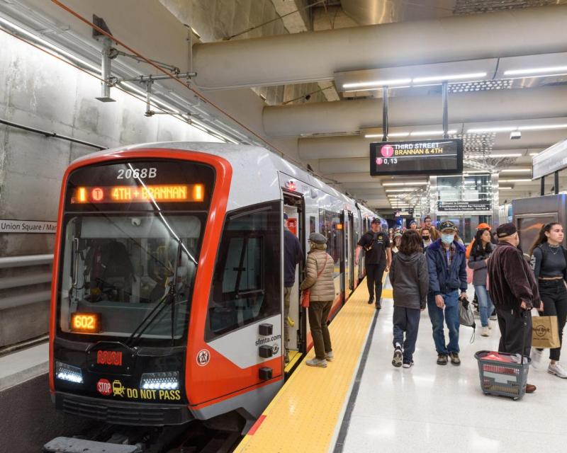 Muni customers boarding the T Third train as it arrives at the station, people walking around.