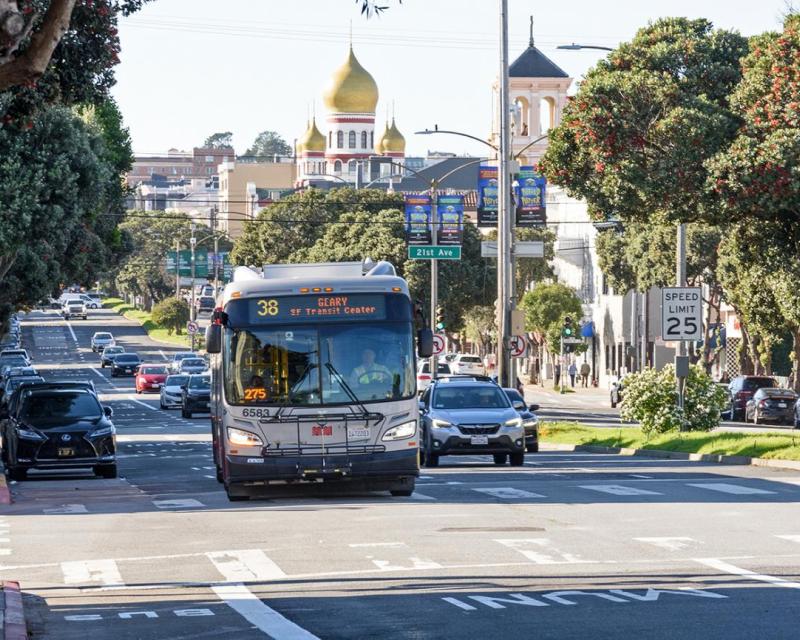 A 38 Geary bus heads east on Geary Boulevard, passing 21st Ave. in a designated transit lane. Cars follow in adjacent lanes.