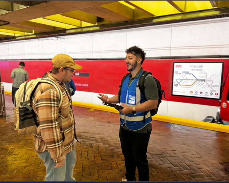 Survey taker wearing a blue vest and badge speaks with a Muni rider on the Powell Station platform for the onboard ridership sur