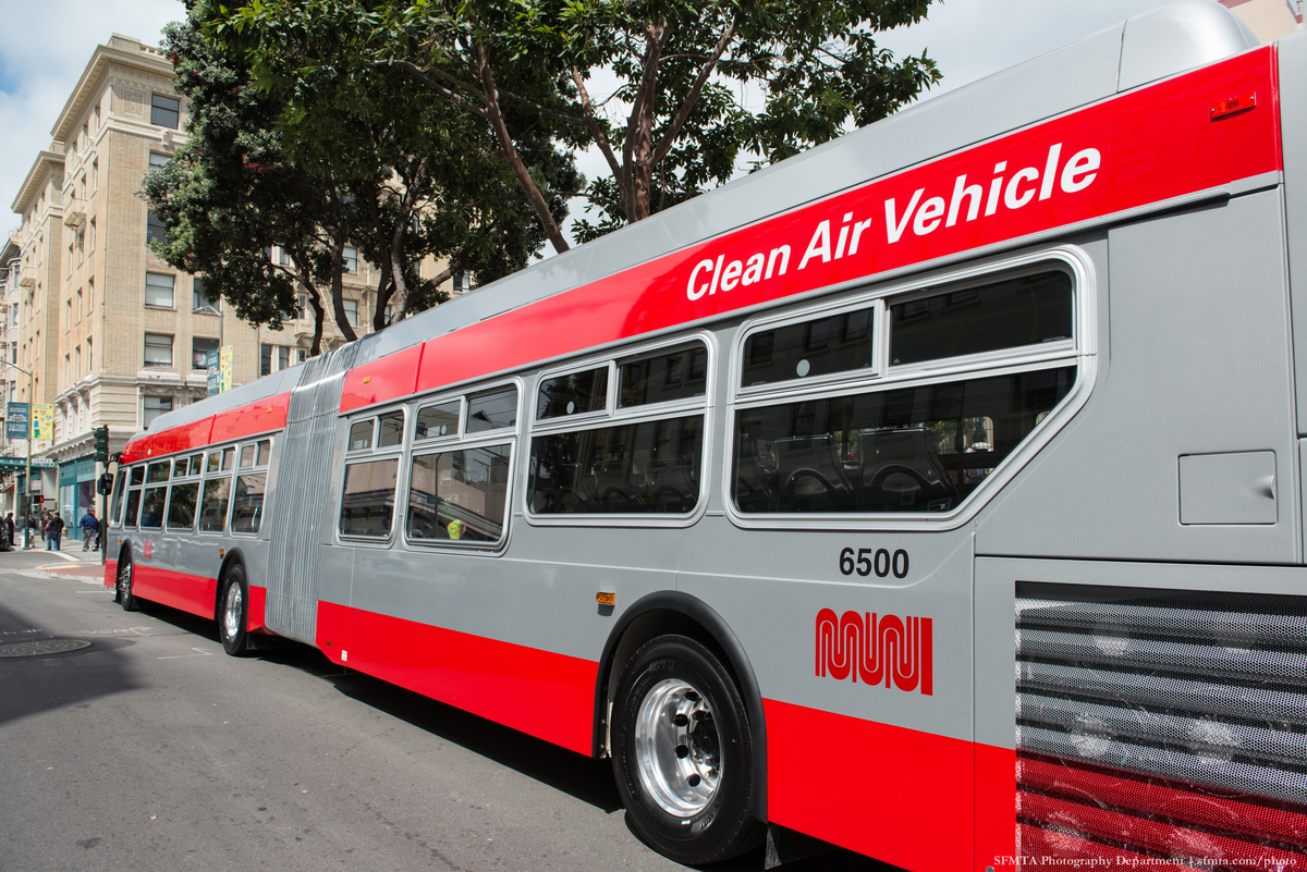 A new, long Muni bus stopped at a street curb. The words "Clean Air Vehicle" are painted on the side.