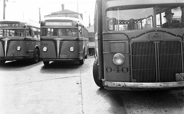 Three vintage 1939 buses are parked in a Muni bus yard. The buses are small and round, with distinctive headlights reminiscent of 1960s Volkswagen buses. In the 1930s these buses were painted with an orange and black color scheme like the Giants baseball team today.