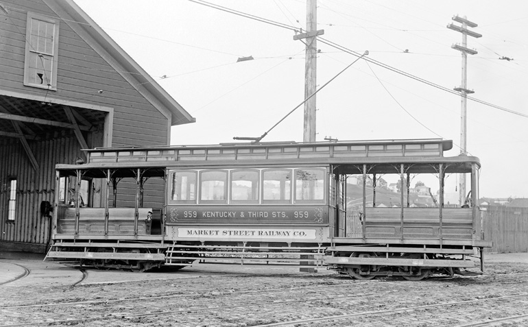 A wooden streetcar is posed for a picture outside of a barn-like building on 3rd Street at 23rd in today’s Dogpatch neighborhood. The streetcar is mostly made of wood, and more closely resembles a cable car than any other type of vehicle. It has open air seconds on either end of the car with benches, and the bars for passengers to grip are ornate. On the side of the car, hand painted lettering reads Kentucky and Third Streets. Unlike a cable car, the streetcar is attached to an overhead power line by a pole that conducts electrical power to the streetcar’s motor.