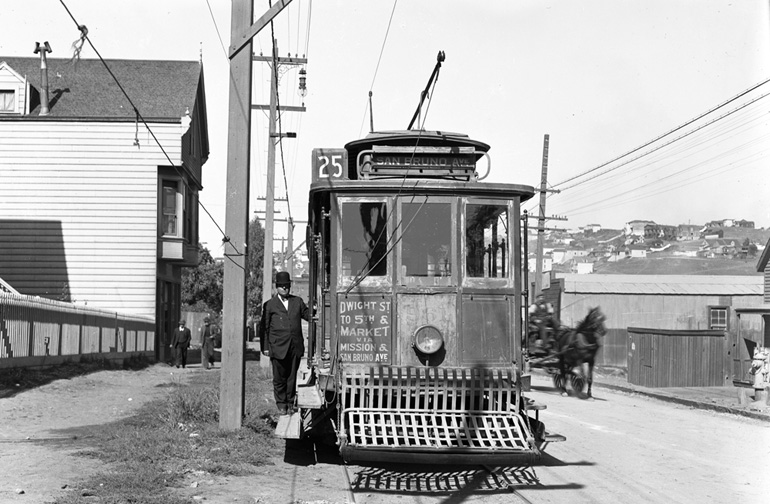 A small, old fashioned streetcar with a sign declaring its destination as San Bruno Avenue is stopped on the tracks with a horse drawn carriage passing on the right. A man in a bowler hat stands on the running board. The year is 1911.