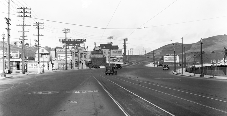 A view looking north on Bayshore Boulevard near Arleta Avenue in 1933. The road is paved and there are streetcar tracks with a waiting area marked by small bumps in the road surface. The Bayshore neighborhood has noticeably grown, with new gas stations and cafes added since the previous image from 1911.