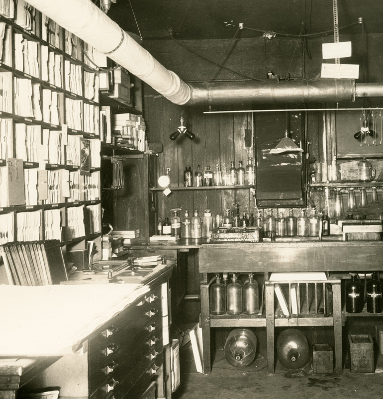 A view of a photography laboratory almost one hundred years ago.  This image was taken around the 1920s and shows thousands of envelopes containing film negatives in shelves on the left side of the lab.  The lab also contains many old fashioned bottles for photography chemicals and an assortment of tongs and clamps for drying prints.