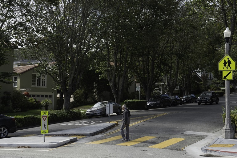 A man walks in a marked crosswalk that features a concrete pedestrian island which narrows the roadway for oncoming drivers. A sign on the island instructs to drivers to yield to pedestrians.