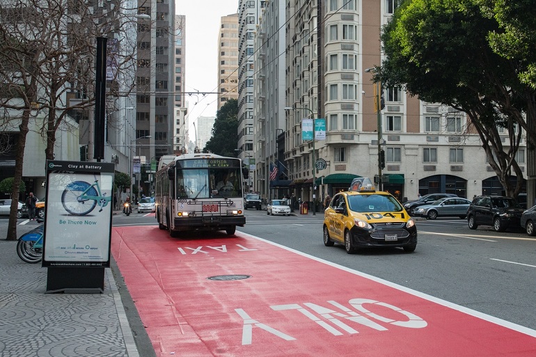 A Muni trolley bus travels down a red-painted transit-only lane on Clay Street in the Financial District alongside a taxi. A bike-share station is on the sidewalk.