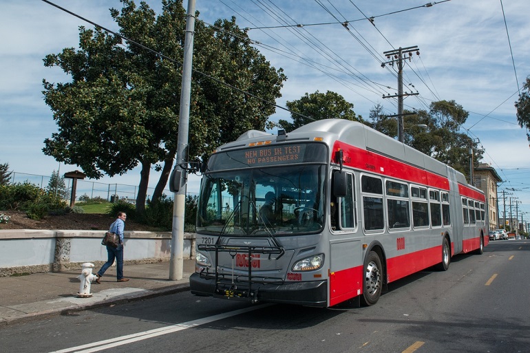 A new 60-foot Muni bus sits curbside with a digital headsign displaying the text, "Bus in test - no passengers." A man on the sidewalk looks at the bus as he walks by."