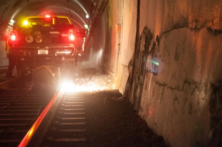 Sparks fly in the Sunset Tunnel as a kneeled crew member cuts the rails behind a stopped work truck. A fellow crew member stands by.