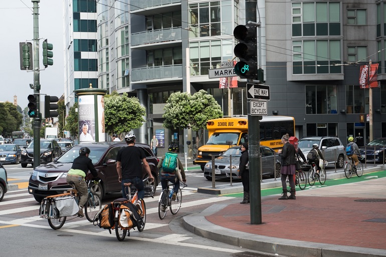 Bike commuters enter a green, curbside bike lane separated from opposing vehicle traffic by a concrete divider and fence.