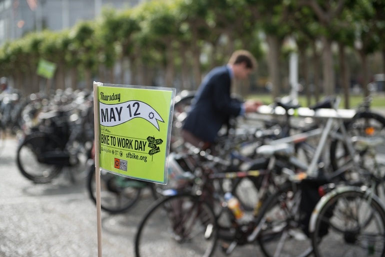 Bikes parked on Civic Center Plaza. In the foreground, a card affixed to a stick to raise above a bike reads, “Thursday, May 12, Bike to Work Day.”]