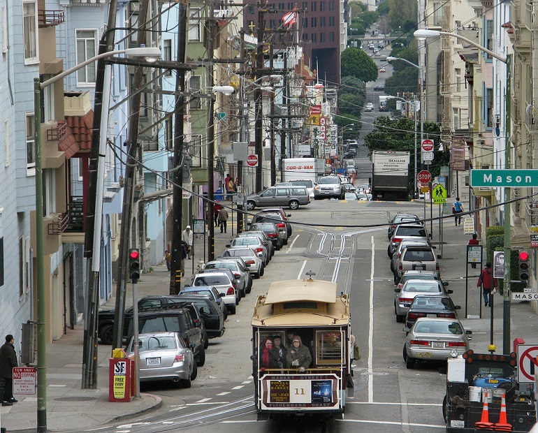 A downhill view of Washington Street at Mason Street. A crowded cable car travels down the street between two rows of parked cars as cars and delivery trucks travel by in the background.