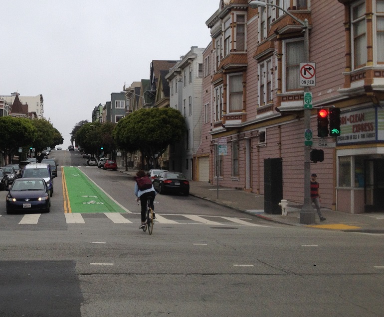 A bicycle rider crosses Oak at Scott Street towards a green left-turn bike lane while. A bicycle signal shows green while a vehicle traffic signal shows red.