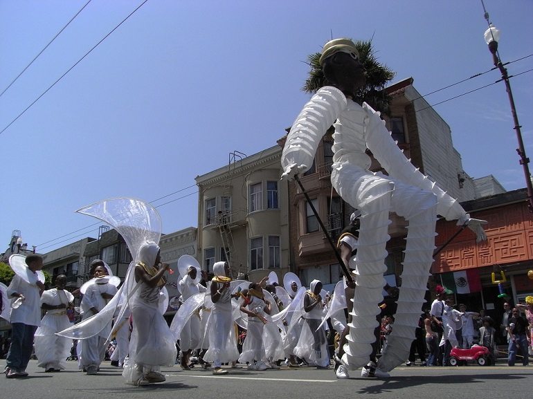 People in costumes parading down the street under a bright blue sky.