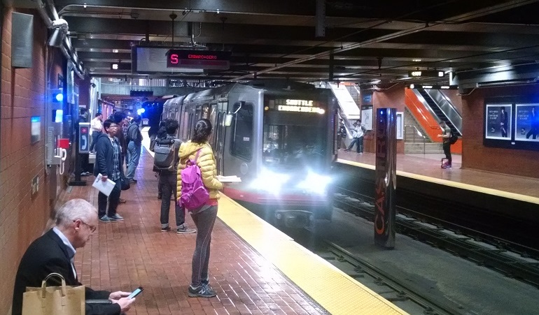 Customers stand on a subway platform while the gray and red Muni train pulls into the station.