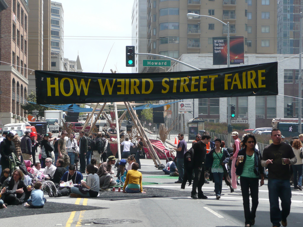 Early afternoon as people gather, sit, and walk around the entrance of a street festival.