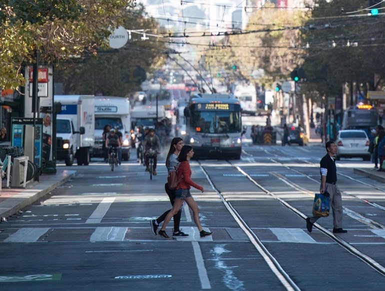 Pedestrians cross Market Street with bicyclists and transit vehicles in the background.