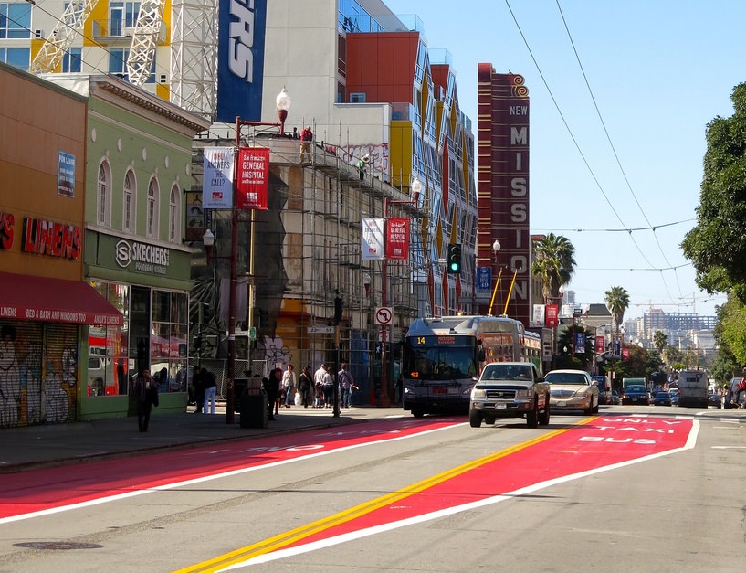 Mission Street at 23rd Street, with a Muni 14 Mission trolley bus traveling down a red lane marked for buses and taxis only, with cars driving in the adjacent lane.