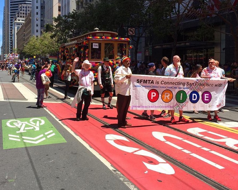 The SFMTA's Pride parade contingent walks down Market Street with a decorated cable car and a banner that reads, "SFMTA is Connecting San Francisco with Pride."