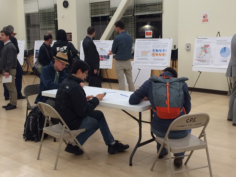 In a large room, people seating at a table filling out a questionnaire while others in the background stand and review graphs and charts on poster boards.