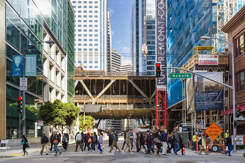 A view of a crowded crosswalk looking south down Fremont Street at Howard Street, with the Transbay Transit Center under construction above.