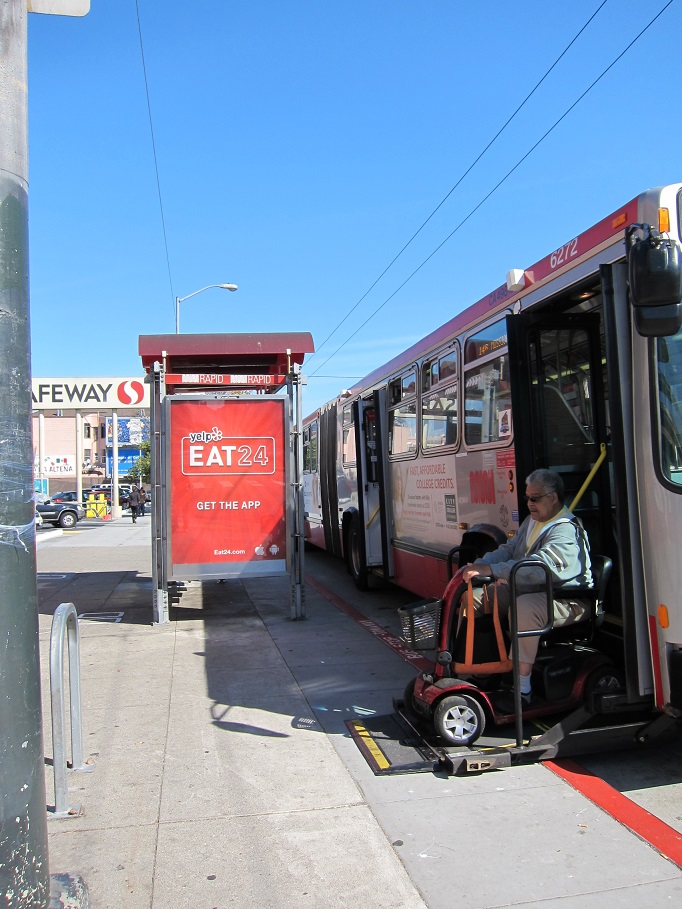 A man in a wheelchair alights from a Muni bus wheelchair ramp on to a transit bulb curb at Mission and 30th streets