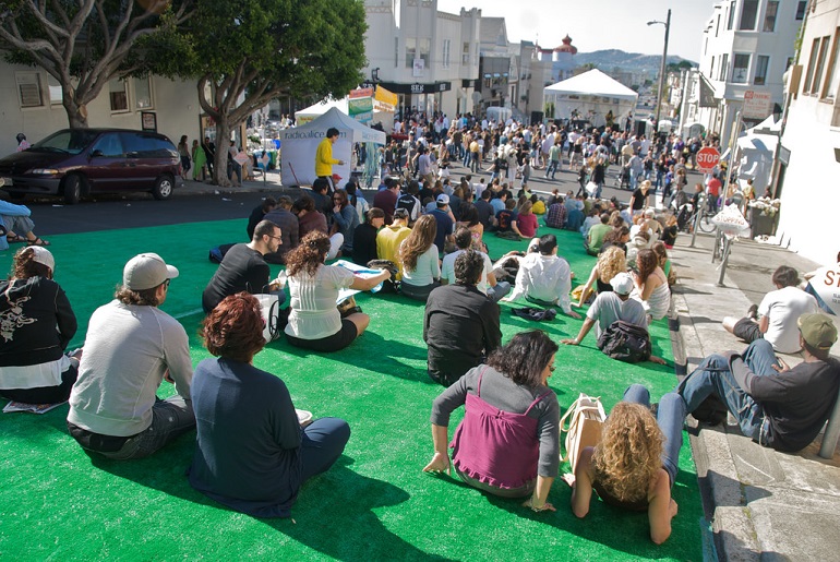 People seating on a green carpet on the street looking at a music stage on a sunny and clear afternoon.