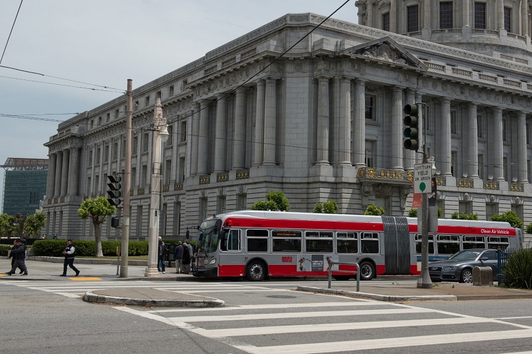 A 60-foot Muni bus stopped on Van Ness Avenue at McAllister Street in front of City Hall.