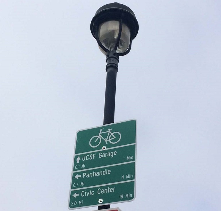 A bike wayfinding sign affixed to a street light pole lists three destinations - UCSF Garage, Panhandle and Civic Center - along with their respective info.