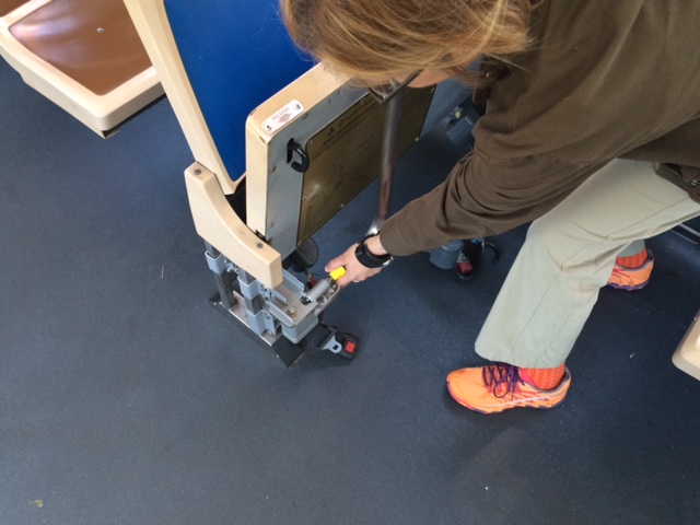 With bus seats flipped up, a woman reaches for a yellow knob at the bottom of a rotational stanchion, which is turned towards the flipped seats and away from the wheelchair space.