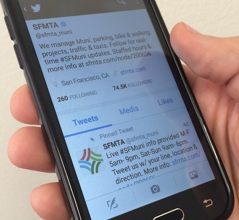 A handheld smartphone features the SFMTA's Twitter page on its screen.