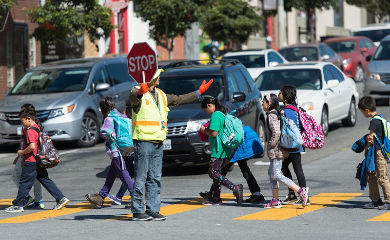 A crossing guard holds a stop sign as children walk in a crosswalk in front of stopped cars.