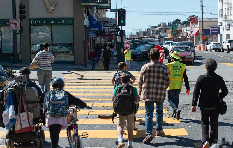 Children and adults cross a street, including a crossing guard and a girl walking a bicycle.