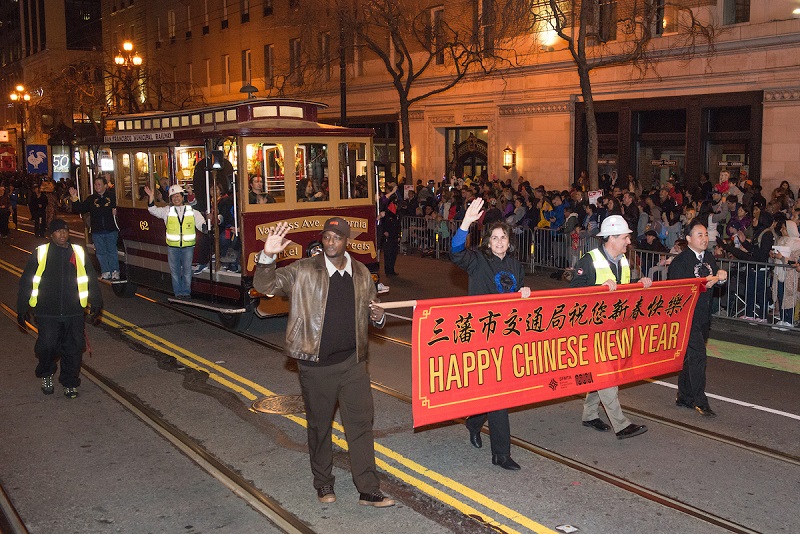 Motorized cable cars glowing with Chinese lanterns is led down Market Street by SFMTA staff carrying a "Happy New Year" banner.