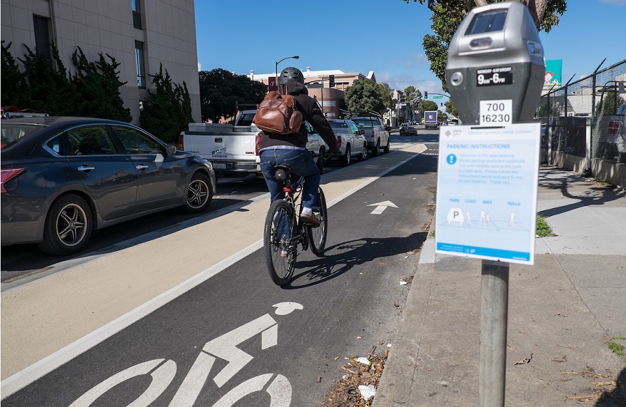 A person bikes on Valencia Street in a bike lane placed between a lane of parked cars and a sidewalk curb, with parking meters featuring instructional flyers.