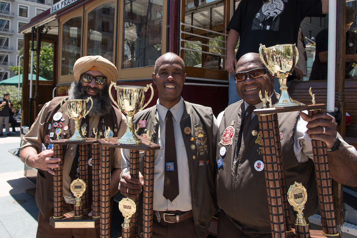 Three Muni Cable Car operators standing in front of the motorized cable car in Union Square with their first, second and third place trophies.