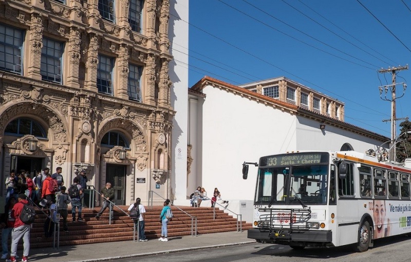 White and orange Muni trolley bus sits in front of Mission High School while students mingle on the steps.