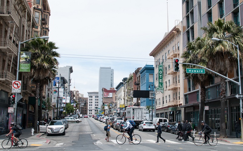 A view of 6th Street as it exists today, with four traffic lanes and people walking and biking across the street.